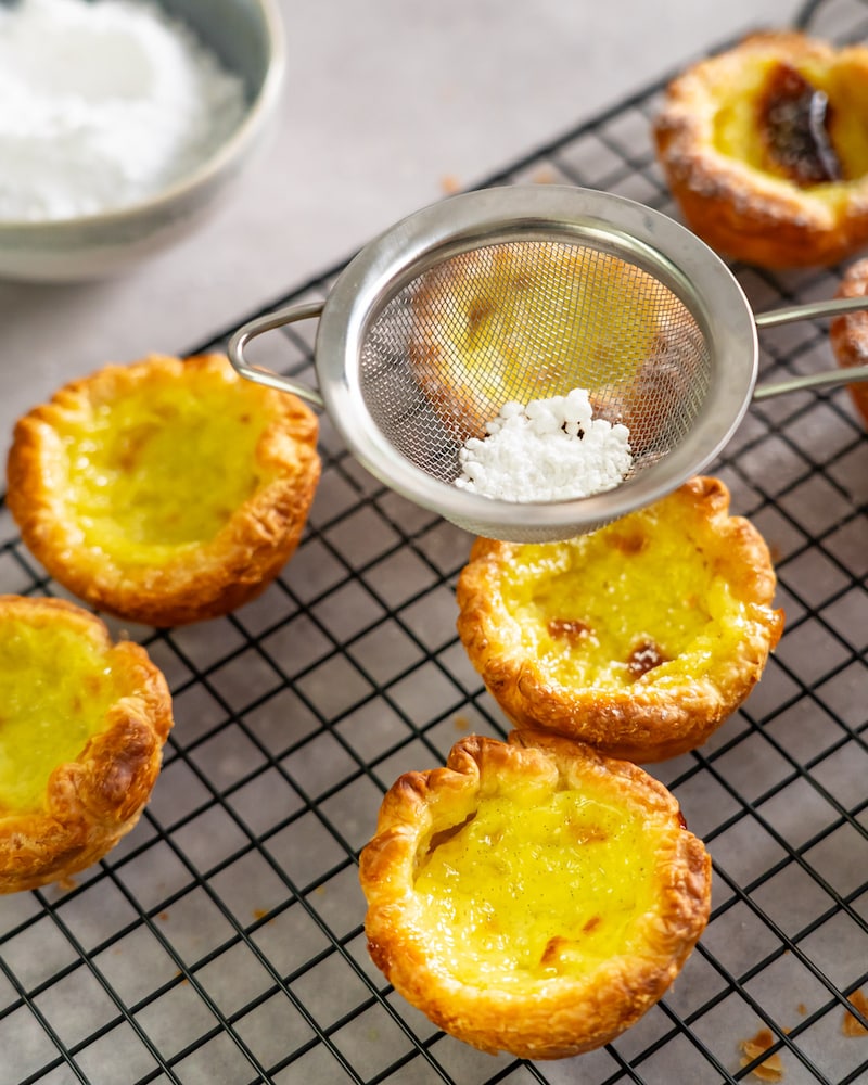 Dieses Bild zeigt wie die fertigen Pastéis de Nata mit Puderzucker bestäubt werden.
