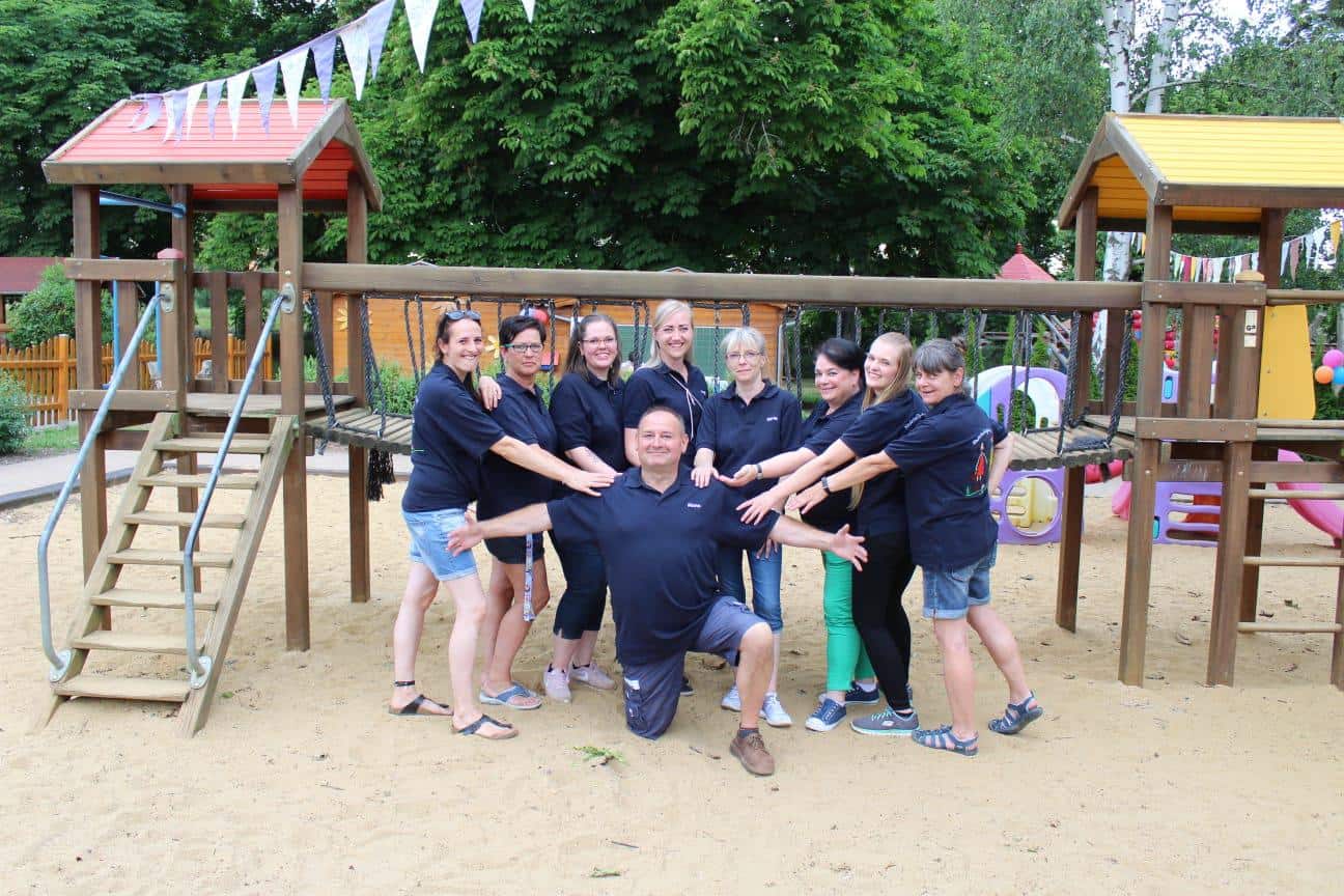 Vereinsmitglieder laecheln froehlich in die Kamera auf dem Spielplatz der Kita