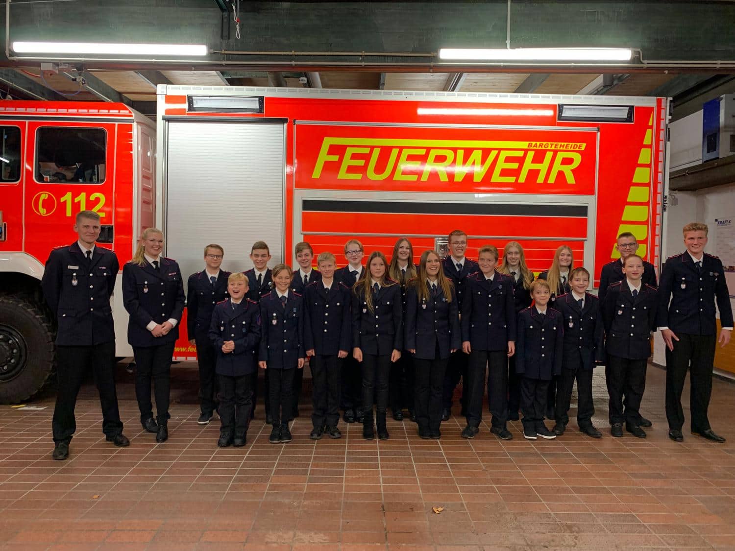 Jugendliche und Vereinsmitglieder stehen vor einem Feuerwehrauto und laecheln in die Kamera