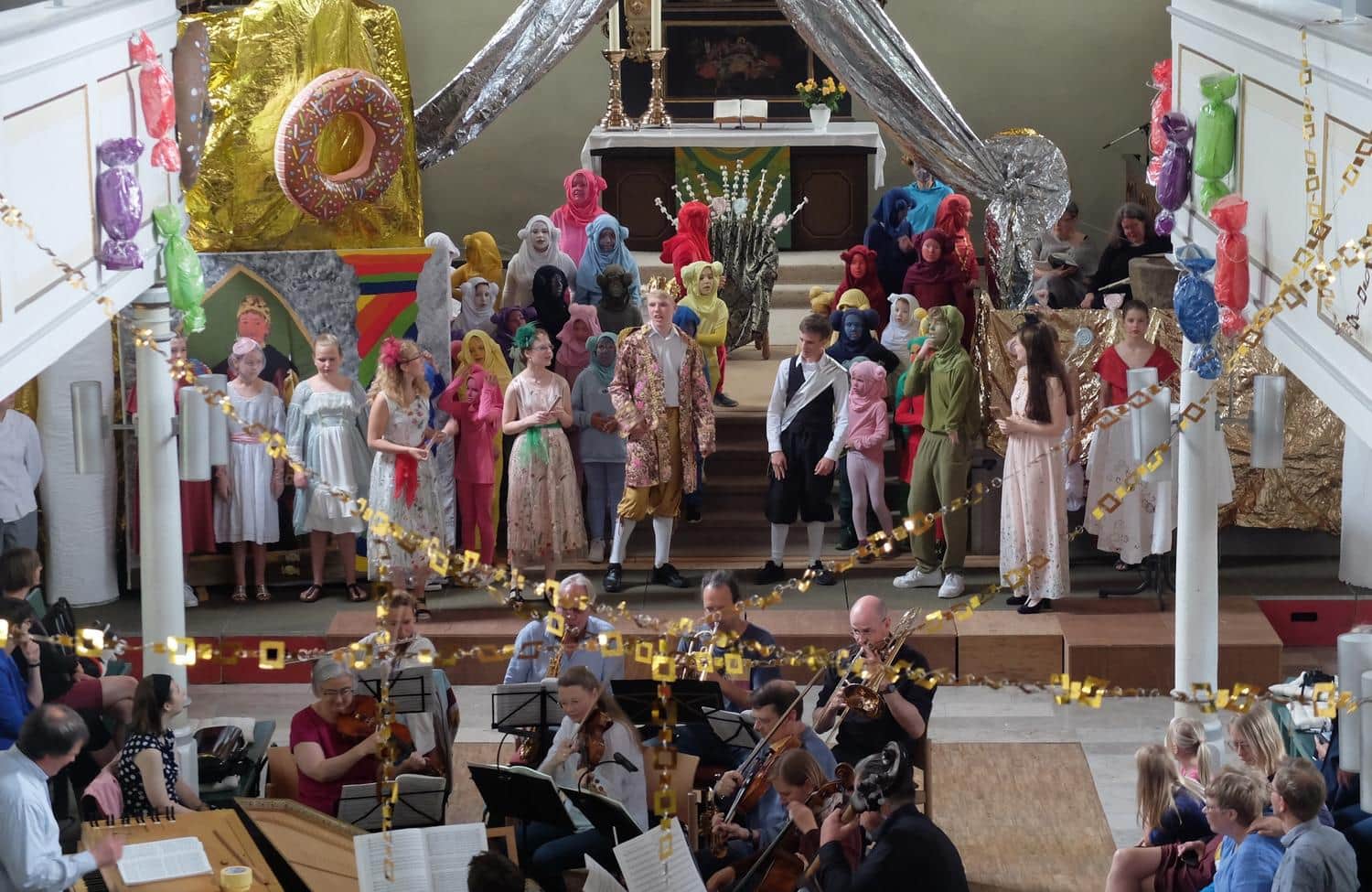Kostuemierte Kinder und Jugendliche spielen ein Stueck auf der Buehne und werden musikalisch begleitet von einem Orchester
