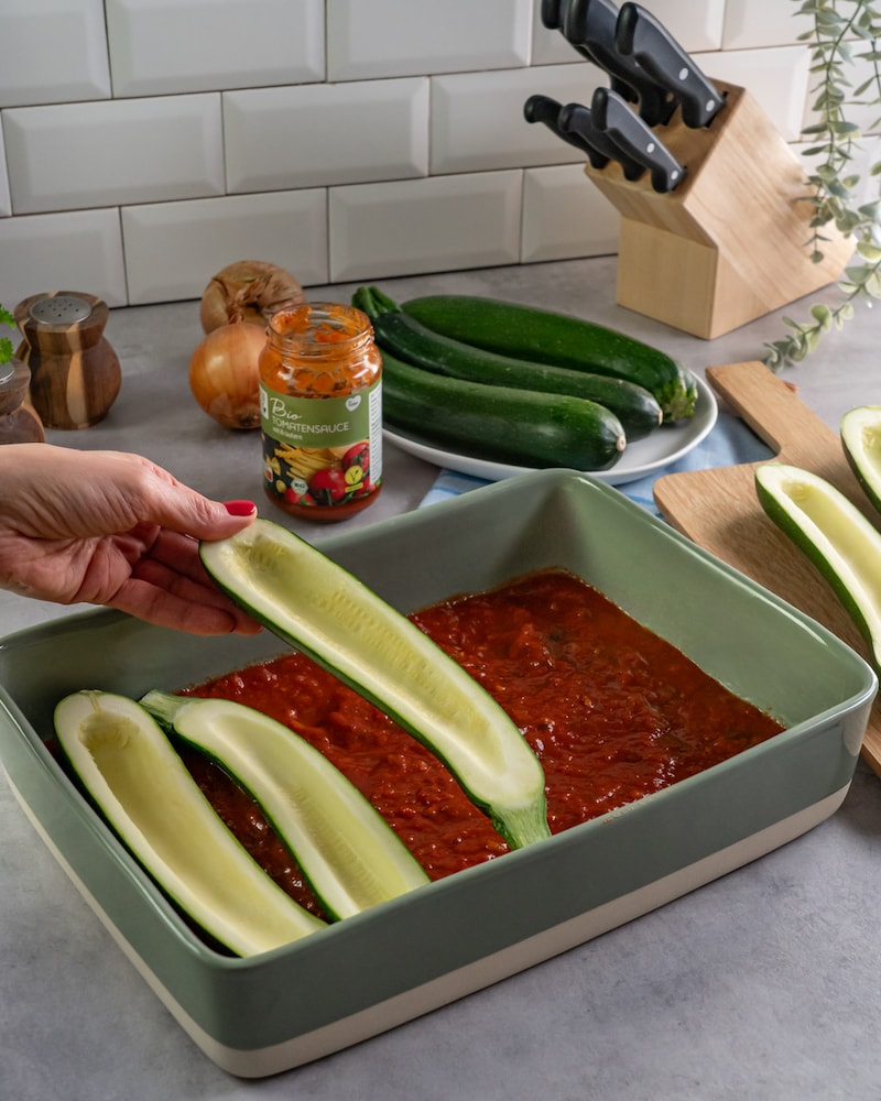 Das Bild zeigt wie die Zucchini in eine Auflaufform mit Tomatensoße gegeben werden.