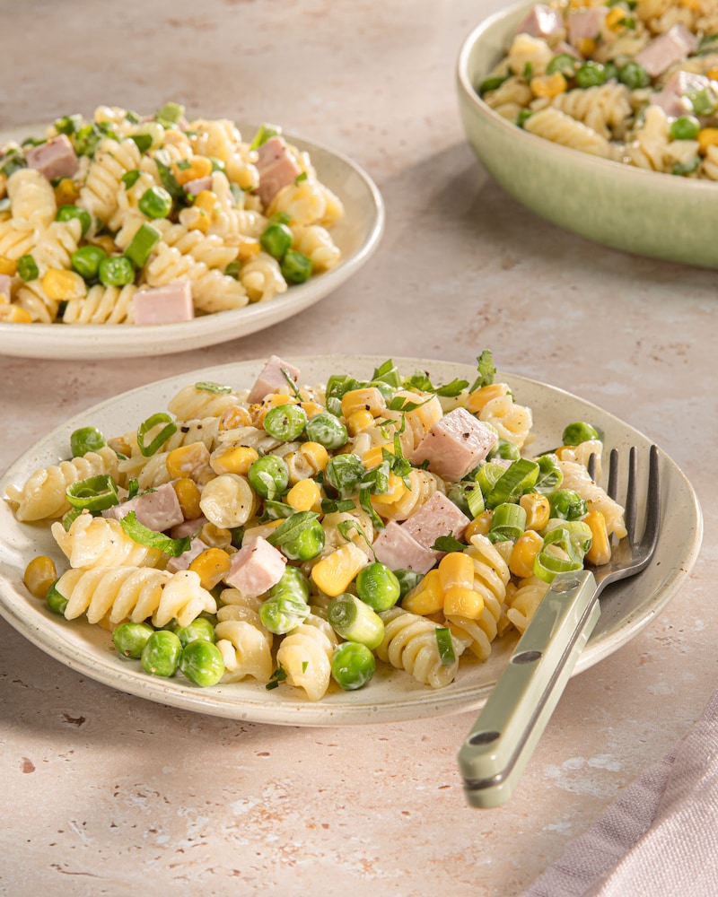 Das Bild zeigt einen Nudelsalat mit Würstchen, Erbsen, Mais und einer Mayo-Soße. 