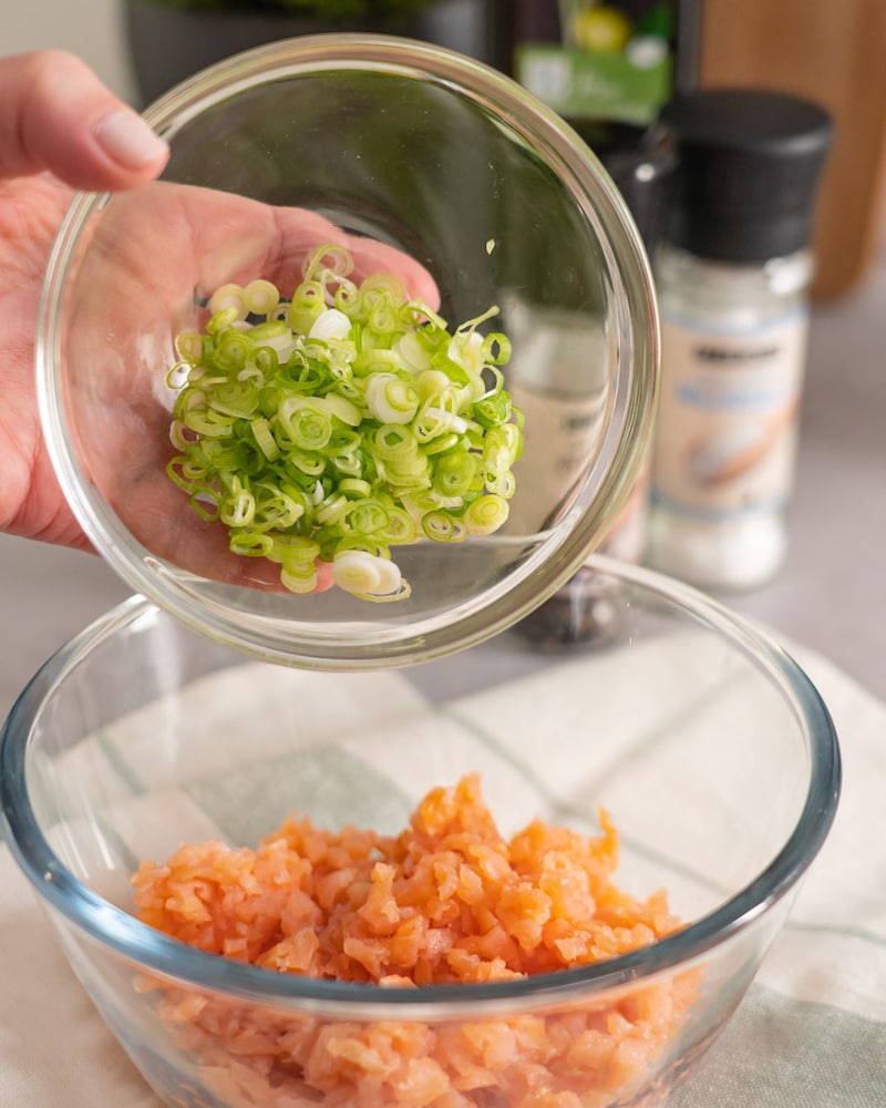 Dieses Bild zeigt wie die geschnittenen Frühlingszwiebeln in eine Schüssel mit Lachs getan werden.