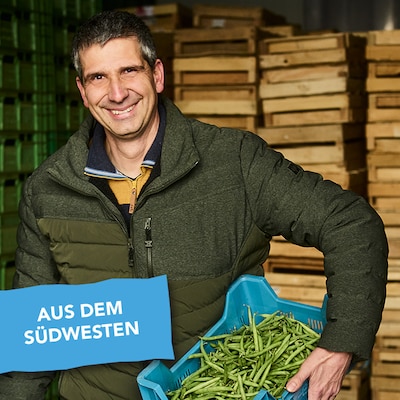 Michael Schmidt trägt lächelnd eine Kiste mit frisch geernteten Bohnen.