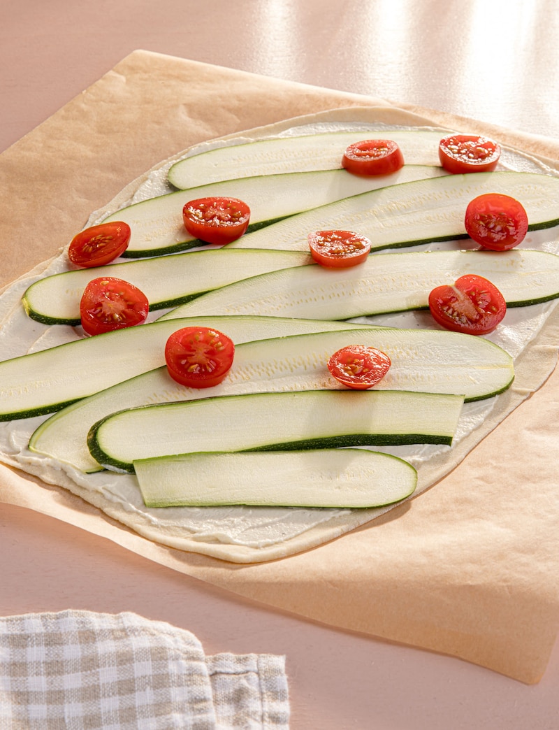 Dieses Bild zeigt wie der Flammkuchen mit Zucchini und Tomaten belegt wird.