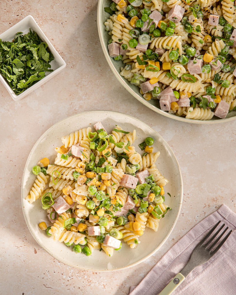 Das Bild zeigt einen Nudelsalat mit Würstchen, Erbsen, Mais und einer Mayo-Soße. 