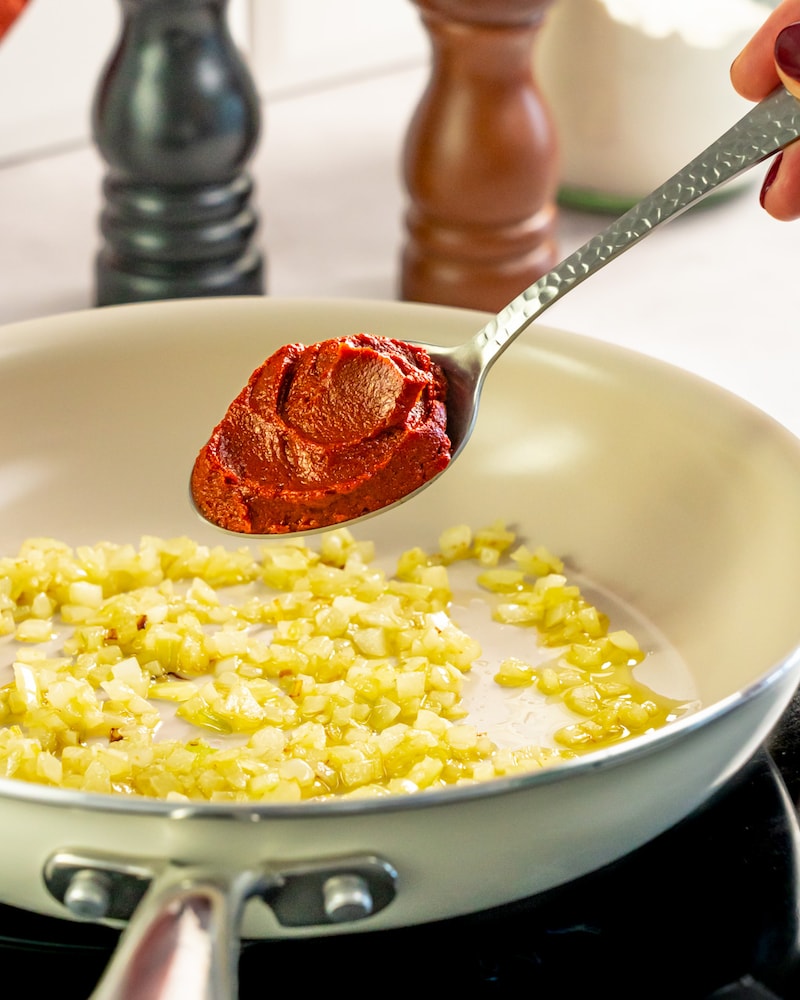 Dieses Bild zeigt wie Tomatenmark in eine Pfanne mit Zwiebeln gegeben wird. 