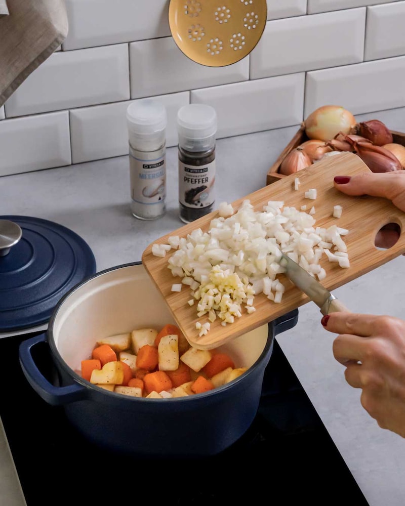 Diese Bild zeigt wie Zwiebeln und Knoblauch zu dem Wurzelgemüse im Topf gegeben werden.