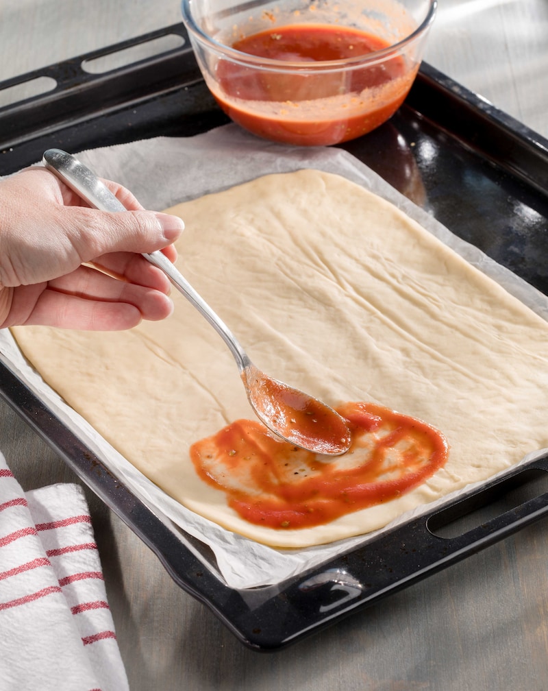 Das Bild zeigt wie Pizzateig mit Tomatensoße bestrichen wird.