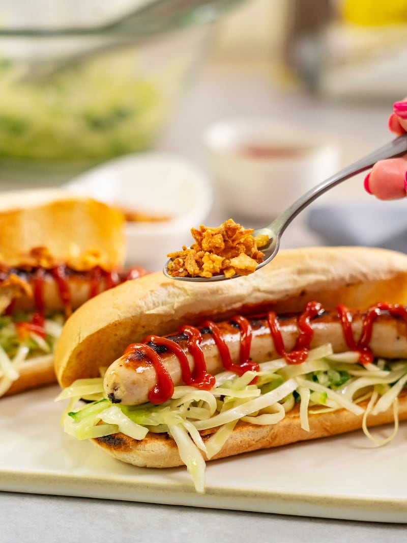 Dieses Bild zeigt wie der Hot Dog vom Grill mit Röstzwiebeln bestreut wird.
