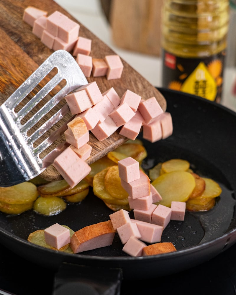 Auf diesem Bild siehst du wie wie der Leberkäse dazugegeben wird.