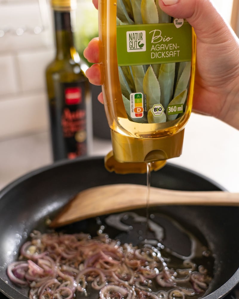 Dieses Bild zeigt wie rote Zwiebeln in einer schwarzen Pfanne angebraten und Agavendicksaft hinzugefügt wird.